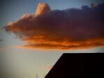 A vibrant sunset showcases a large, dramatic cloud painted in deep orange and pink hues. The cloud hovers over the silhouette of a building's rooftop, capturing an image perfect for any online art gallery. A small, bright speck in the background hints at an aircraft or star against the evening sky.