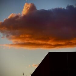 A vibrant sunset showcases a large, dramatic cloud painted in deep orange and pink hues. The cloud hovers over the silhouette of a building's rooftop, capturing an image perfect for any online art gallery. A small, bright speck in the background hints at an aircraft or star against the evening sky.