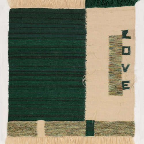 a woven textile features a deep green section on the left and a cream section on the right. the word “love” is spelled vertically in green on the cream side. cream fringe borders the top and bottom edges.