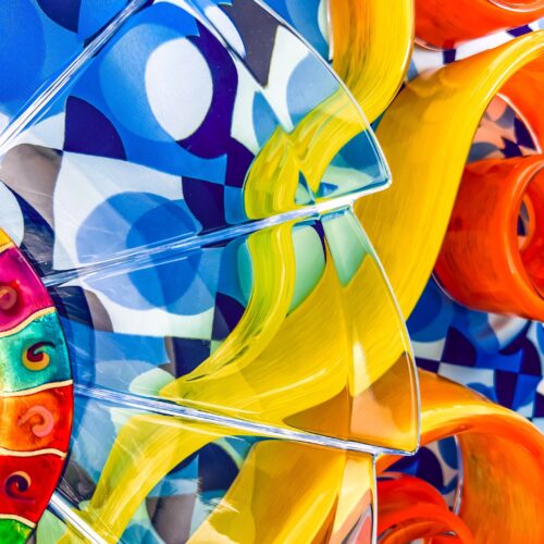 abstract close up of a colorful glass sculpture featuring swirling yellow, orange, and red shapes against a vibrant blue and white geometric background.