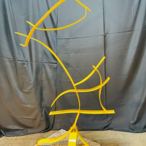 abstract yellow metal sculpture with curving, intersecting lines standing on a speckled base, set against a gray draped fabric backdrop and concrete floor.
