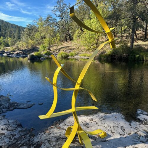 a bright yellow abstract metal sculpture stands on a rocky bank beside a calm river, surrounded by trees and greenery under a clear blue sky.
