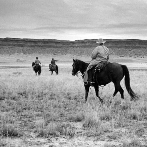 three people on horseback ride across a grassy plain under a cloudy sky, with rocky hills in the distance. the nearest rider is in the foreground, wearing a wide brimmed hat and jacket.