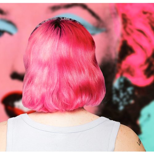 A person with short, bright pink hair seen from behind, positioned in front of a colorful pop-art portrait of Marilyn Monroe.