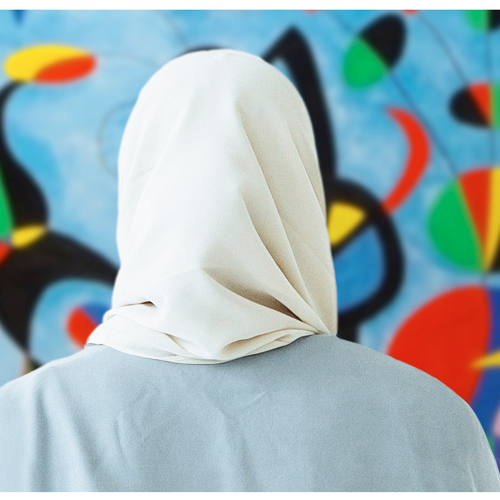 A person wearing a headscarf stands with their back to the camera, looking at a colorful abstract painting.