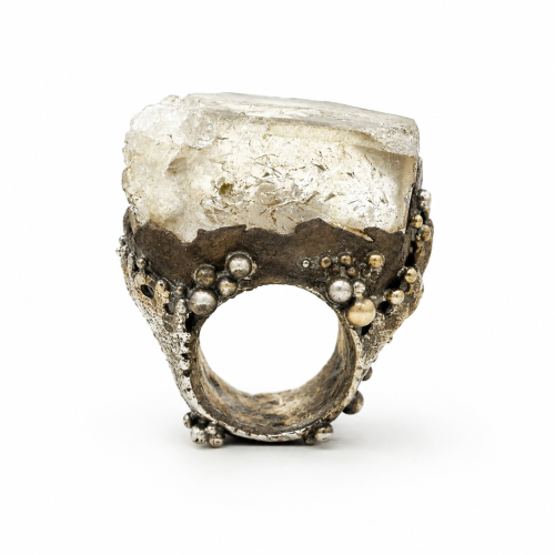 A chunky, textured metal ring featuring a large, rough-cut clear gemstone set in a darkened band decorated with small metallic spheres.