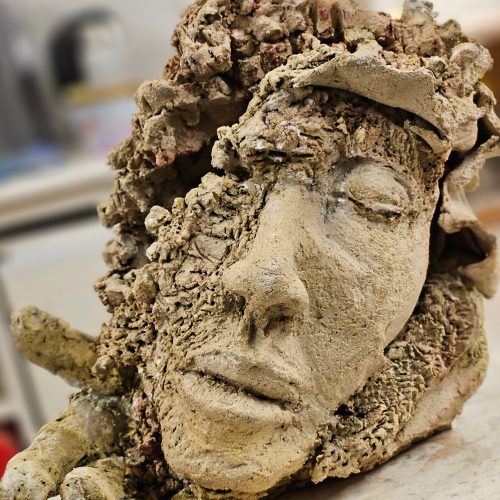 A textured, weathered sculpture of a human face with closed eyes and a serene expression, partially surrounded by rough, stone-like material, rests on a surface in an indoor setting—an evocative piece perfect for adding character to any e-commerce décor collection.