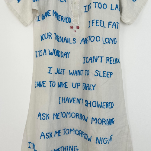 A white short-sleeved dress embroidered with blue text phrases expressing various excuses and frustrations, such as “I have a headache,” “I feel fat,” and “Ask me tomorrow night.”