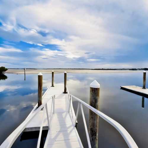 A white floating dock with railings extends into calm, reflective water under a cloudy sky.