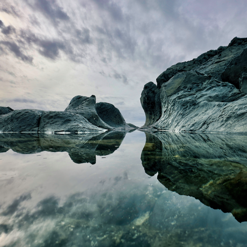 Jagged rock formations reflecting in a still tide pool under a cloudy sky.