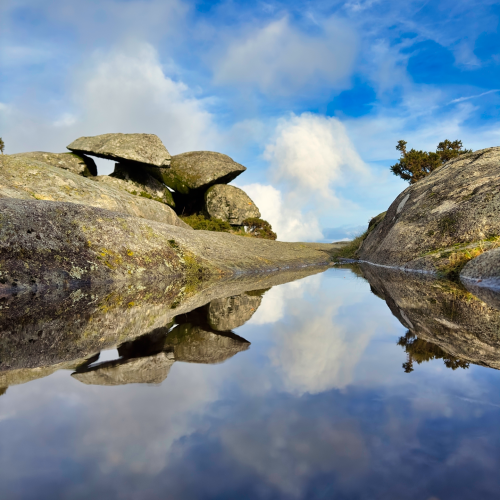 A large rock formation reflected in a small pool of water under a cloudy blue sky.