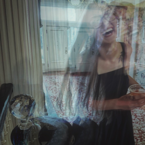 A blurred woman in a black dress laughs while holding a drink, standing in a brightly lit, ornate hallway with floral wallpaper and chandeliers.