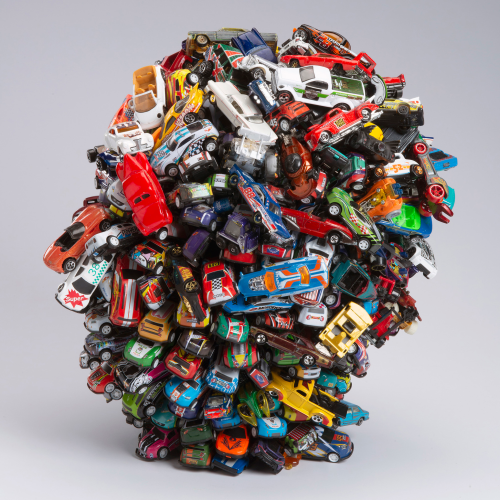 A large, dense sculpture made of numerous assorted toy cars piled together into a spherical shape.