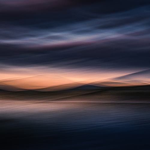 Abstract image of a sunset over water, with blurred, wavy streaks of dark blue, orange, and purple, creating a dreamy, painted effect across the sky and horizon.