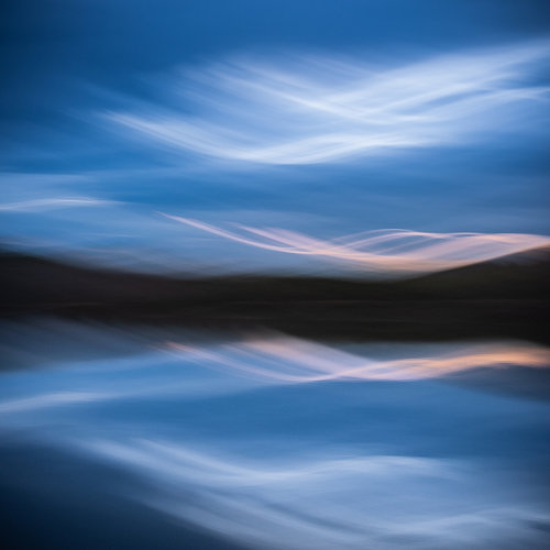 Abstract image of a calm lake reflecting a blue sky with blurred white and pink clouds, creating a dreamy, painterly effect.