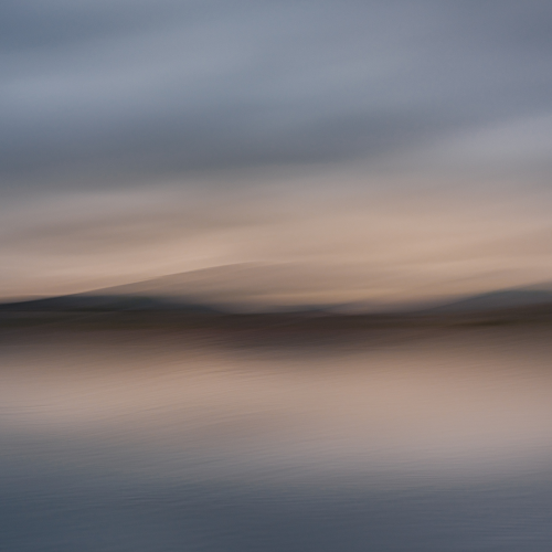Blurry landscape with soft blue, gray, and peach tones. Water reflects an indistinct horizon and cloudy sky, evoking a peaceful, abstract mood.