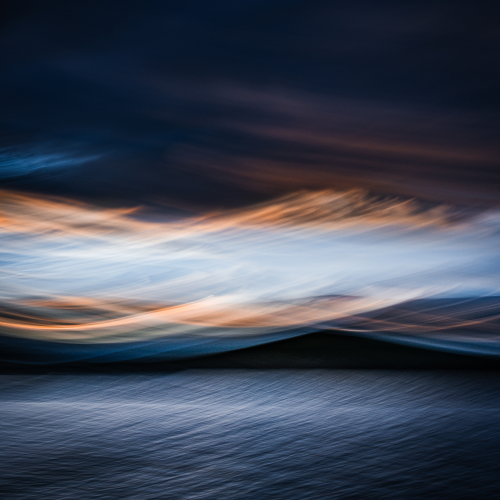 Abstract image of a tranquil body of water and a dark hill under a dramatic sky with streaked clouds in shades of blue, orange, and pink at sunset or sunrise.