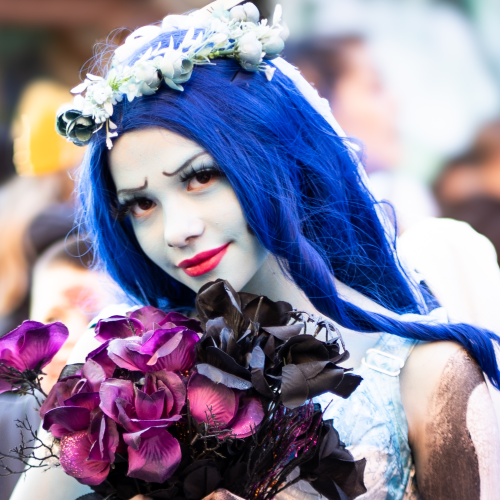 A person in a Corpse Bride costume with blue hair, a floral headpiece, and pale makeup, holding a bouquet of purple and black roses.