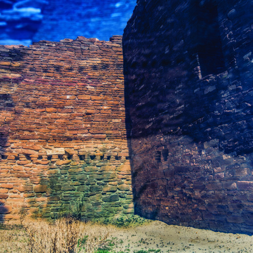 stone ruins with tall, weathered walls stand under a bright blue sky. shadows and sunlight fall across the textured stones, and some dry grass grows along the sandy ground inside the structure.
