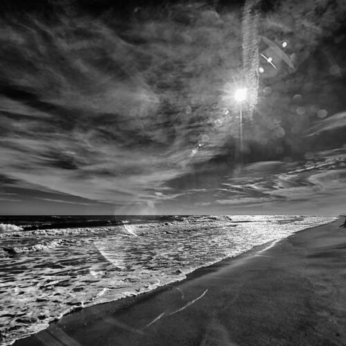 black and white photo of a beach with waves washing onto the shore under a dramatic, cloudy sky. the bright sun is partially obscured by clouds, creating lens flare and reflections on the wet sand.