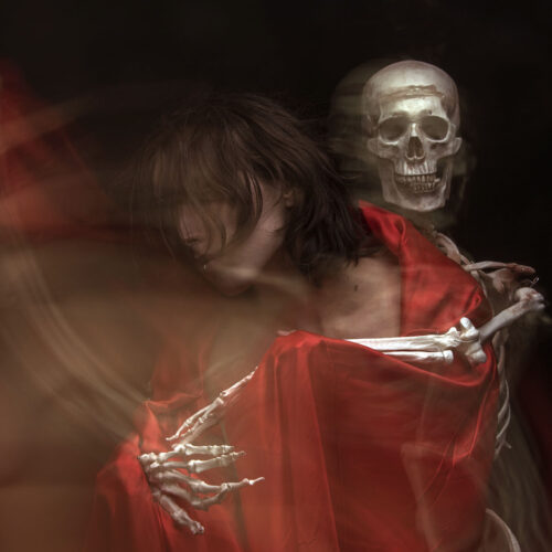 a person draped in a flowing red cloth is embraced from behind by a human skeleton. the image is blurred, creating a sense of motion and an eerie, ghostly atmosphere.