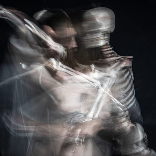 a blurred, double exposure image shows a person intertwined with a skeleton against a dark background, creating a ghostly, surreal effect as their forms partially merge and overlap.