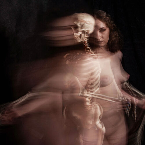 a blurred, artistic photograph of a nude woman standing with a superimposed skeleton image, creating a ghostly, double exposure effect against a dark background.