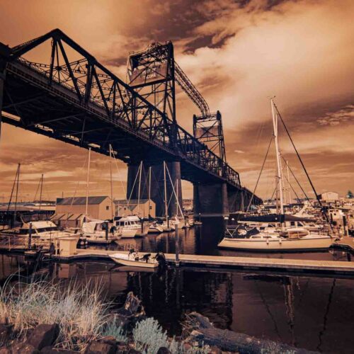 a marina with several docked sailboats sits beneath a large industrial lift bridge. the sky and water have a reddish orange hue, creating a dramatic infrared effect. buildings and boats fill the background.