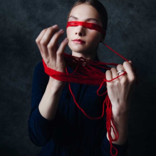 a woman in a dark outfit stands against a dark background, her eyes covered with red yarn. she holds and entangles the yarn with both hands, her expression calm and contemplative.