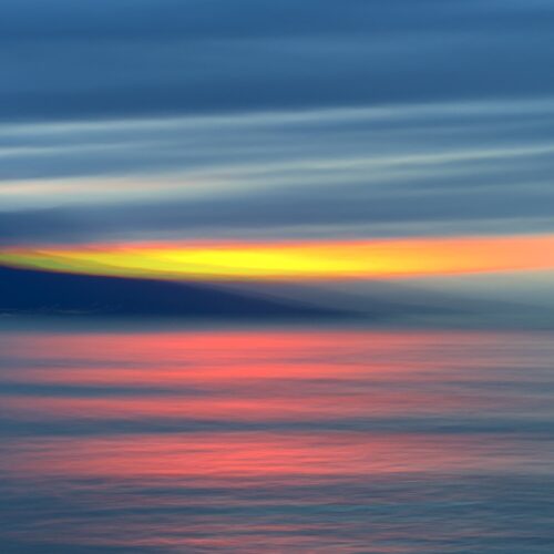 abstract image of a colorful sunset over the ocean, with vibrant streaks of orange, yellow, and pink reflecting on the calm water beneath a dramatic, cloud filled sky.