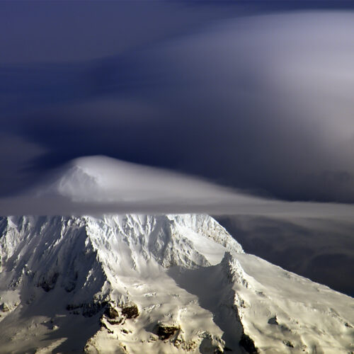 snow covered mountain peak partially obscured by dramatic, smooth, layered lenticular clouds, creating a striking contrast between the bright white snow and the darker, swirling sky.